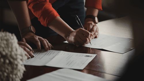 Two people lean over a wooden desk signing documents. 