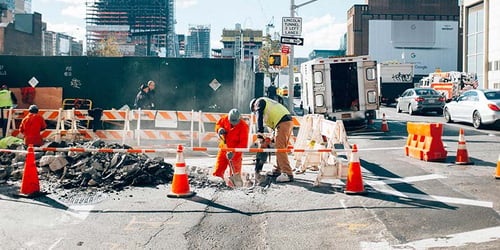 Construction workers are working inside of a hole on the side of the road during city traffic.