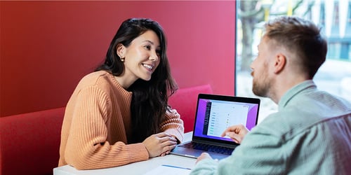 Two people sit across from one another at a table reviewing work on a laptop together.