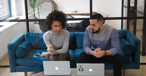 Two people sit beside one another discussing things in front of their laptops.