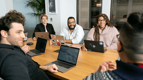 A group of people sit around a conference table having a discussion while on their laptops.