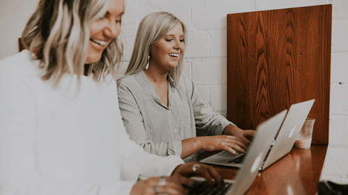 Two women smiling while working on their computer at a joint desk.