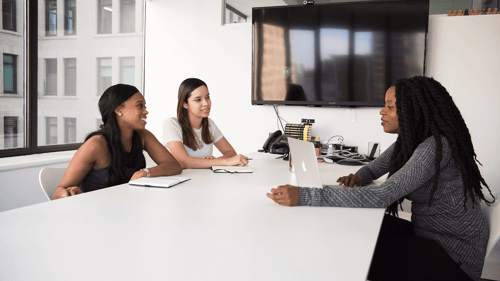 Three people sit around a conference room table having a discussion in an interview like manner. 