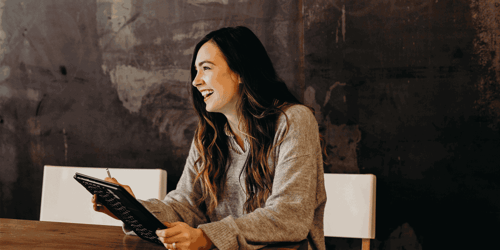 A person sits at a table smiling into the distance while holding a tablet.