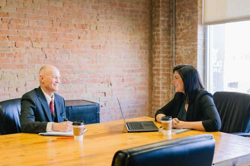 Two people sit across from one another dressed in formal business attire in a conference room