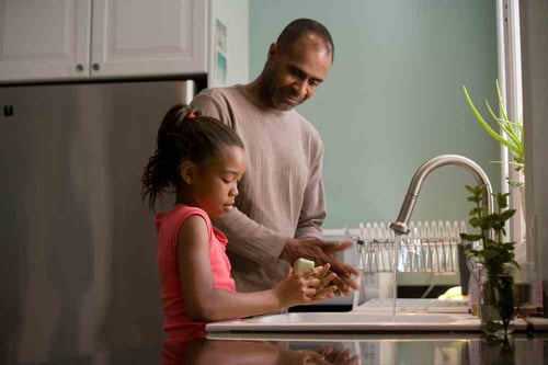 A parent helps wash dishes with their child at the sink