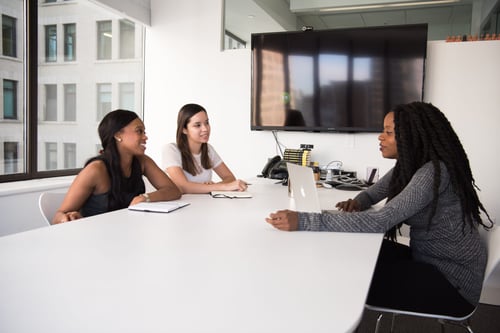 Three people sit in a conference room having a discussion. There is a TV on the wall and a large open window.