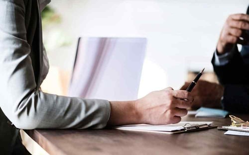 Two people sit at a table having a discussion. One person is prompting the other to sign the paper that is on the table.
