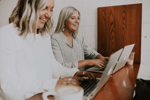 Two people sit at a shared desk laughing and typing on their laptops