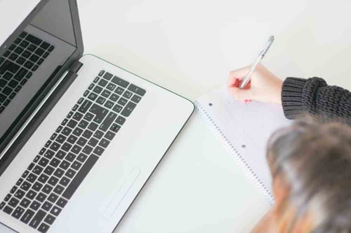 A person sits at their desk taking notes on their pad and paper while also looking at their laptop screen