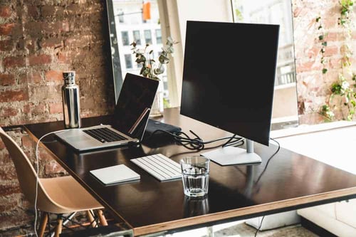 A laptop is resting a wooden desk locate next to a large window with a cityscape view