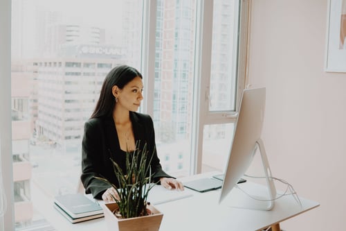 A person sits at their desk in front of a large open window working on their computer 