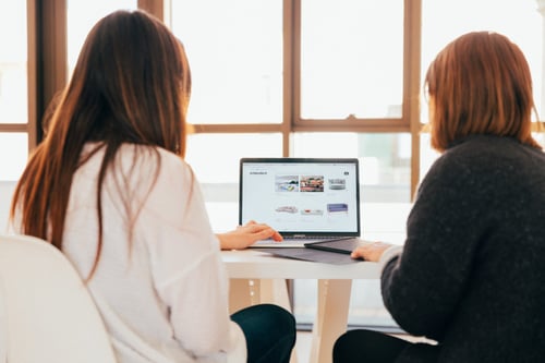 Two people sit in front of a window working on the computer together