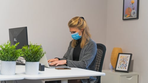 An individual with a face covering on works at their desk on their laptop