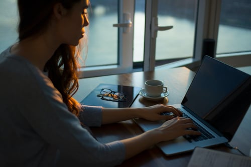 A person types on their laptop at a desk in front of an open glass door