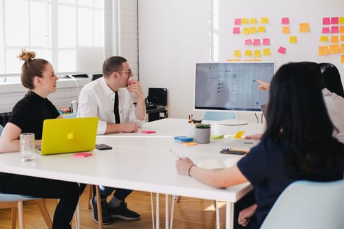 Group of coworkers discussing the presentation on a larger screen. People in the group are taking notes on pads of paper or their laptops.