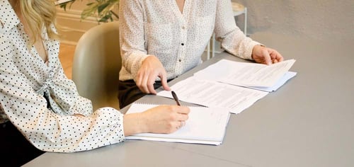 Two people sit at a desk together reviewing paperwork
