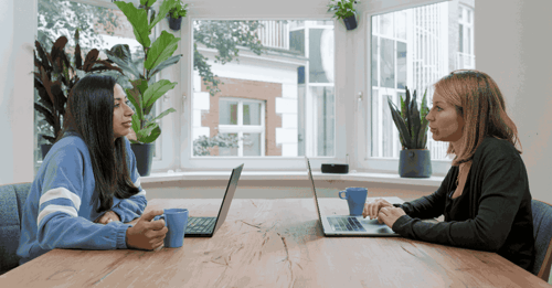 Two people sit on their laptops having a conversation across the table from one another. 