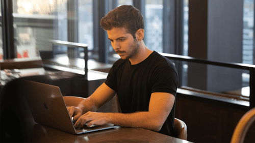 A person sits at their desk typing on their computer in front of large glass windows.