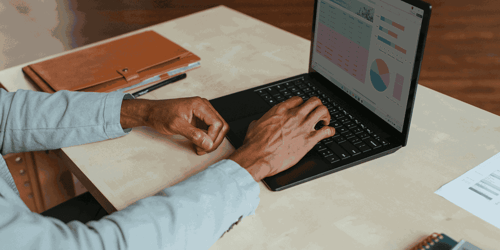 A person sits at a table looking at graphs on their computer with only their hands and computer in view.