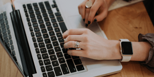 A person wearing an apple watch with their nails painted black types on a computer with only their hands in view.