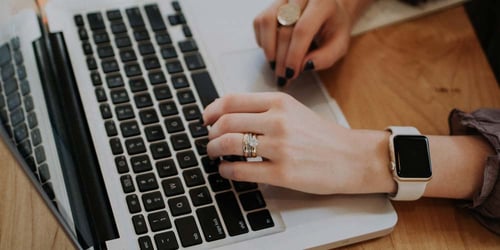 A person wearing an apple watch with their nails painted black types on a computer with only their hands in view.
