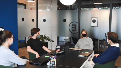 A group of masked individuals are sitting in a conference room having a discussion and working on their laptops