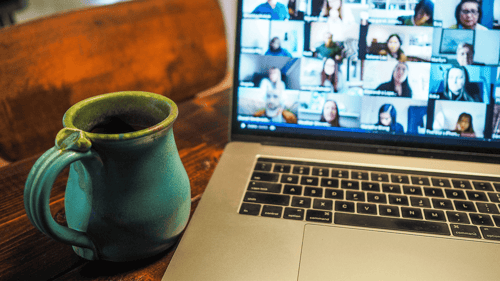 A laptop with a conference call open sits on a wooden table beside a cup of coffee