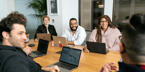 A group of employees sits around a conference room table with their laptops smiling at one another.