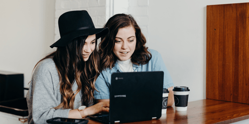 Two young employees sit together reviewing something on a laptop.