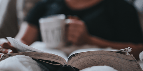 Someone sits reading a book and drinking out of a mug.