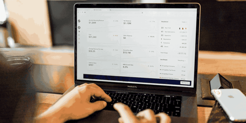 A person with only their hands in view types on their computer with a stock market dashboard on their screen.