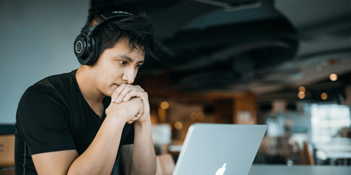 A person with headphones on sits at a desk with their hands rested under their chin viewing content that is out of view on their laptop.