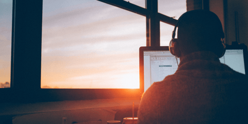 A person sits at their computer next to a window. They have on headphones and seem to focusing intently on their screen.