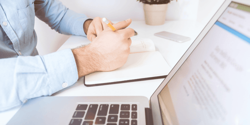 A person sits at their work desk writing in a notebook with their laptop screen in view.