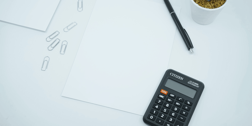 A calculator, piece of paper, a pen, and a few paperclips all sit on a white table.