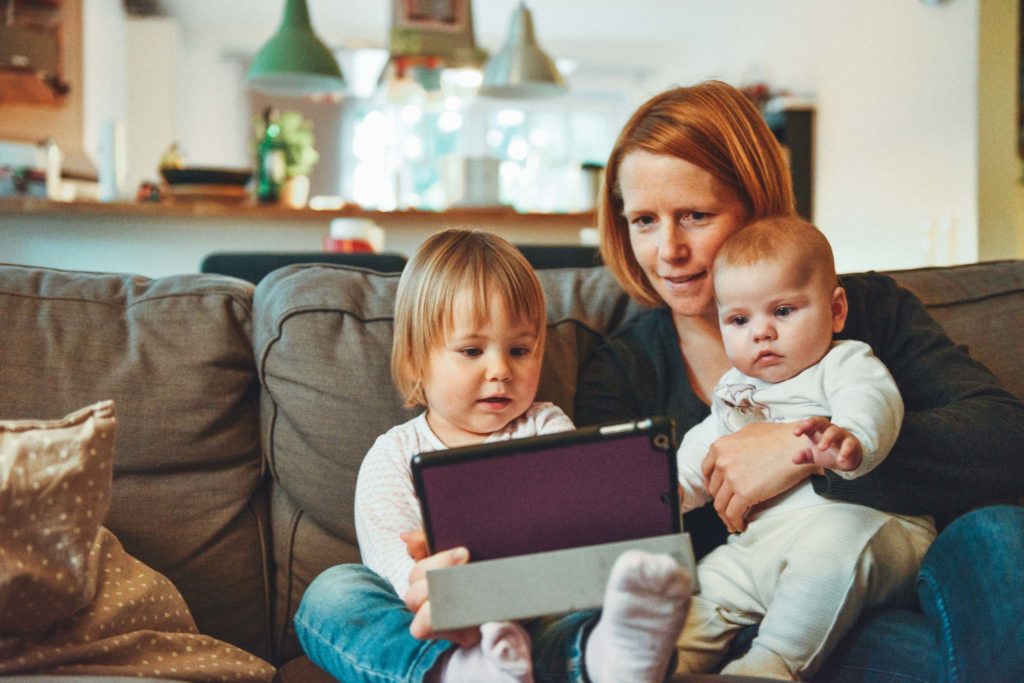 A person sits on the couch looking at a tablet device with their two children