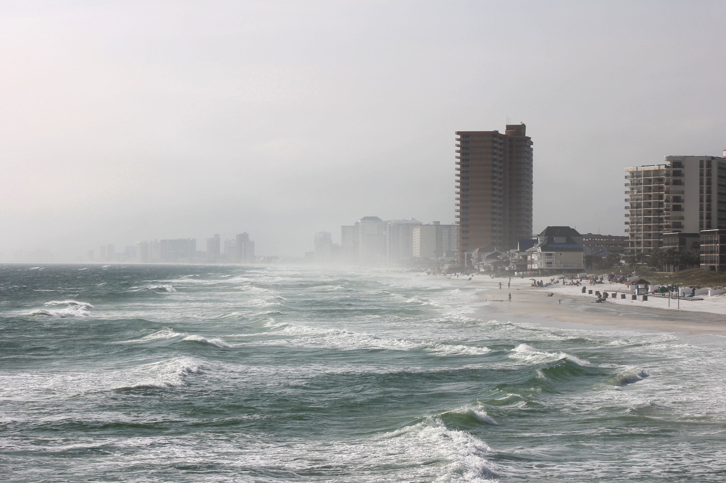 A cityscape alongside a beach setting is met with large crashing waves and gray sky