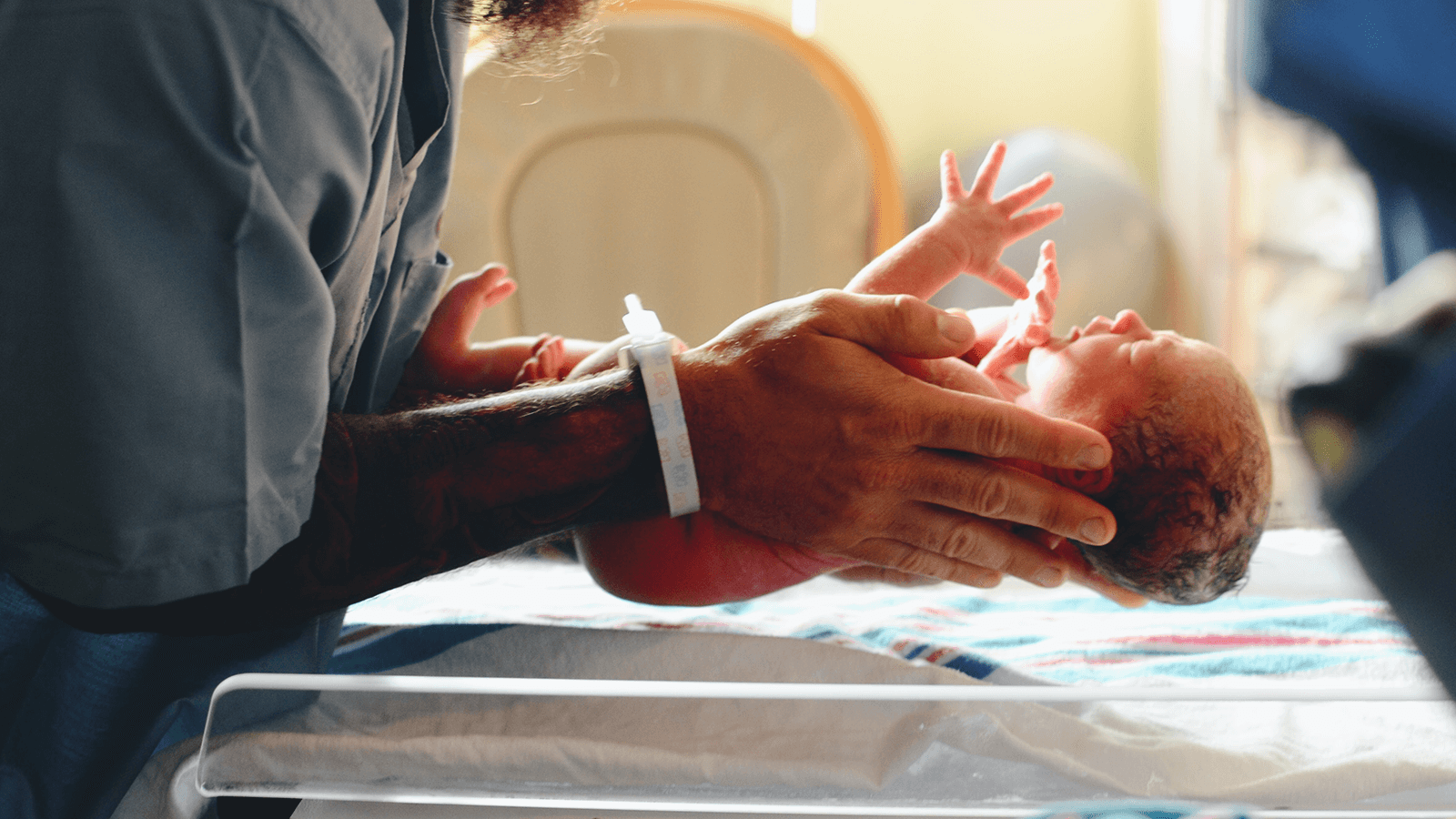 An adult with a hospital bracelet on has their hands in view while holding a newborn baby over a plastic hospital bassinet.