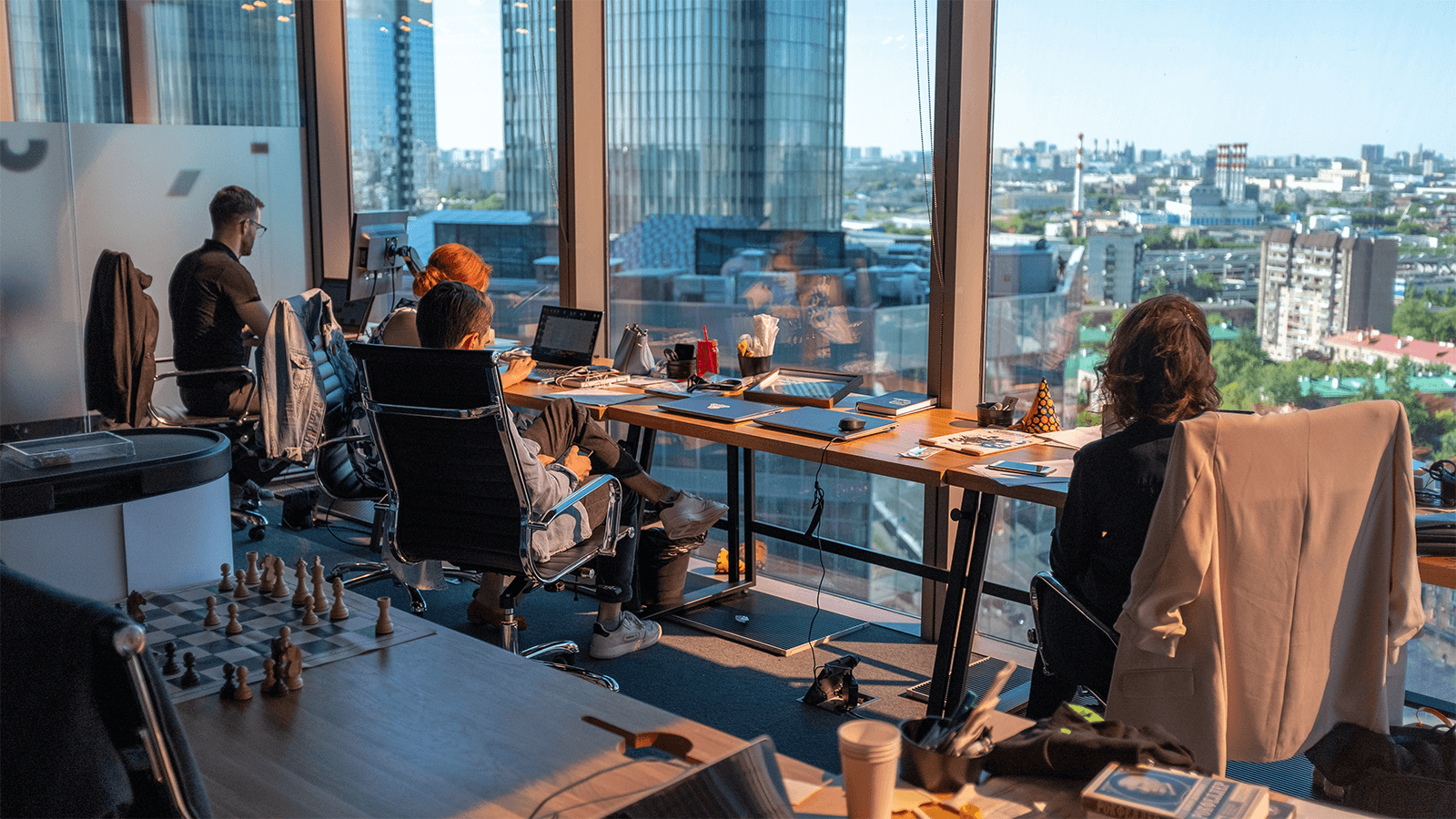 A group of people work at a communal workspace overlooking a cityscape. 