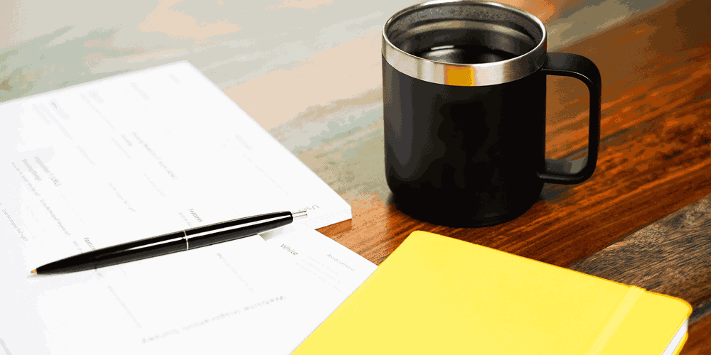 Two notepads, a pen, and a coffee cup all sit on top of a wooden table. 